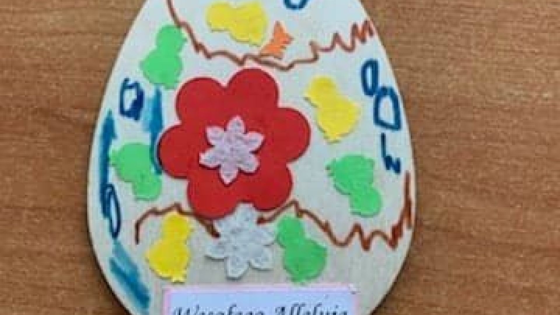 Wielkanocna pisanka z drewna, ozdobiona kolorowymi kwiatami, kurczaczkami i kredkami, z napisem  wesołego Alleluja. Autor: Maciej A.