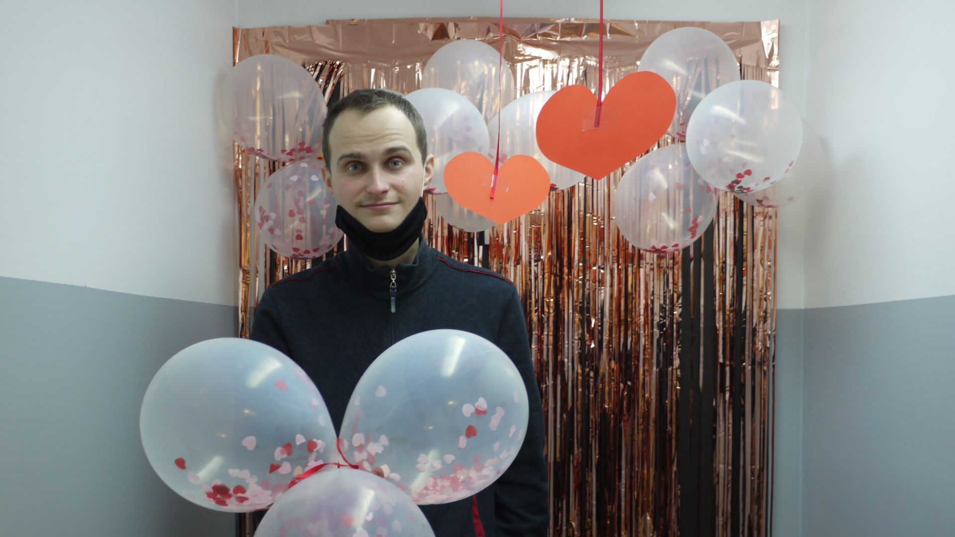 Krzysztof C. trzyma w ręku balony i pozuje na ściance udekorowanej balonami, serpentynami i serduszkami.