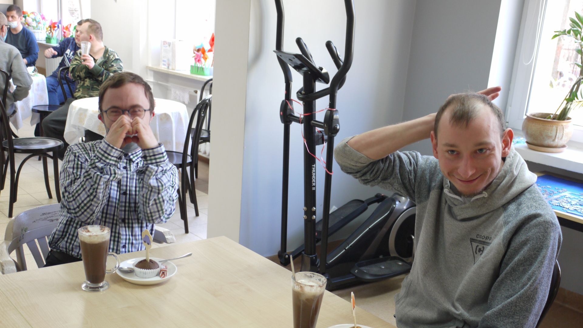 Maciej A. i Jakub K. siedzący przy stoliku z czekoladą na gorąco i babeczką. Jakub K. pokazuje do zdjęcia dłonie ułożone w kształt serca.