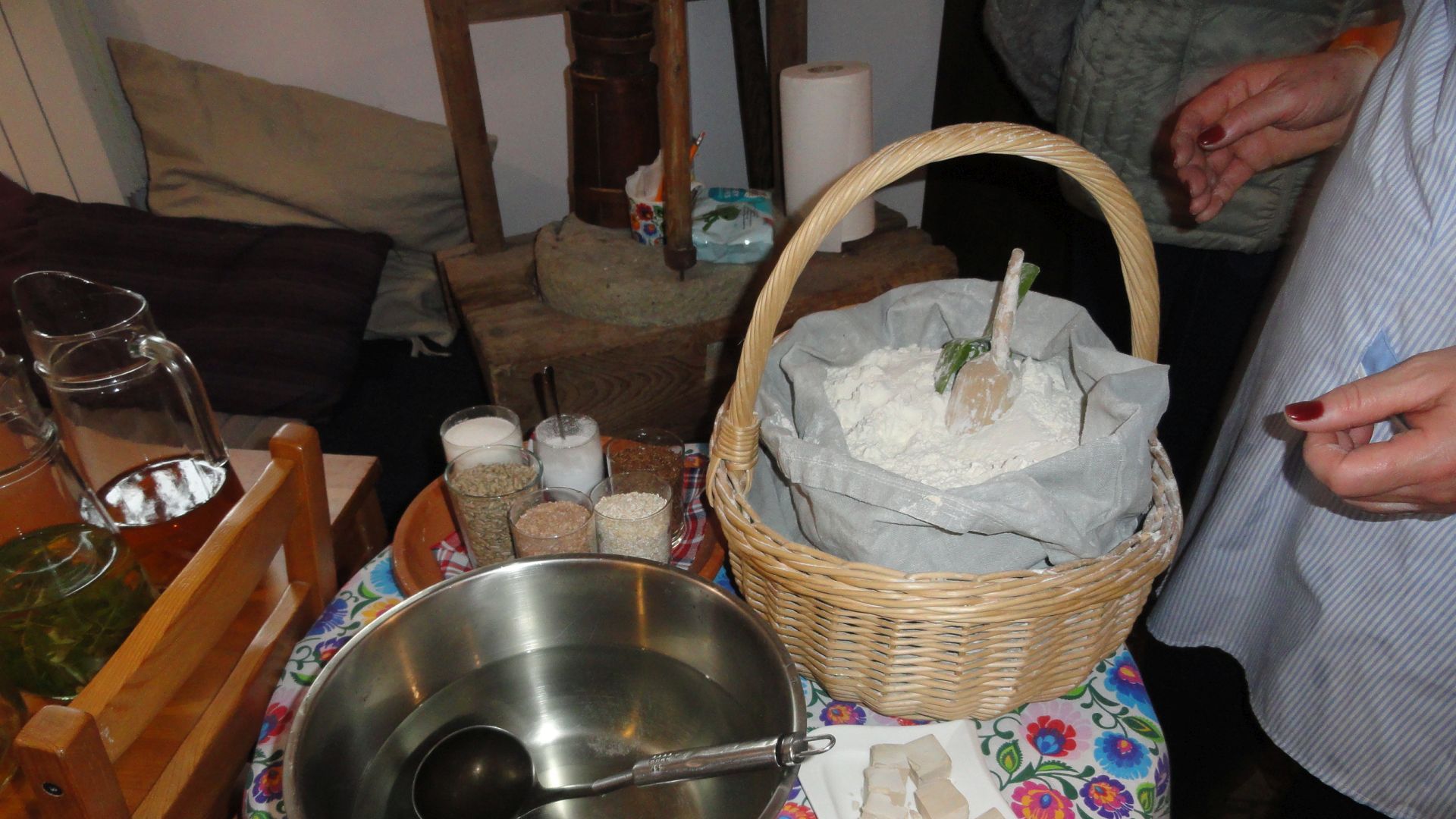 Stolik z produktami potrzebnymi do wypieku chleba: miska z wodą, kosz z workiem mąki, kostki drożdży, szklanki z ziarnami. 