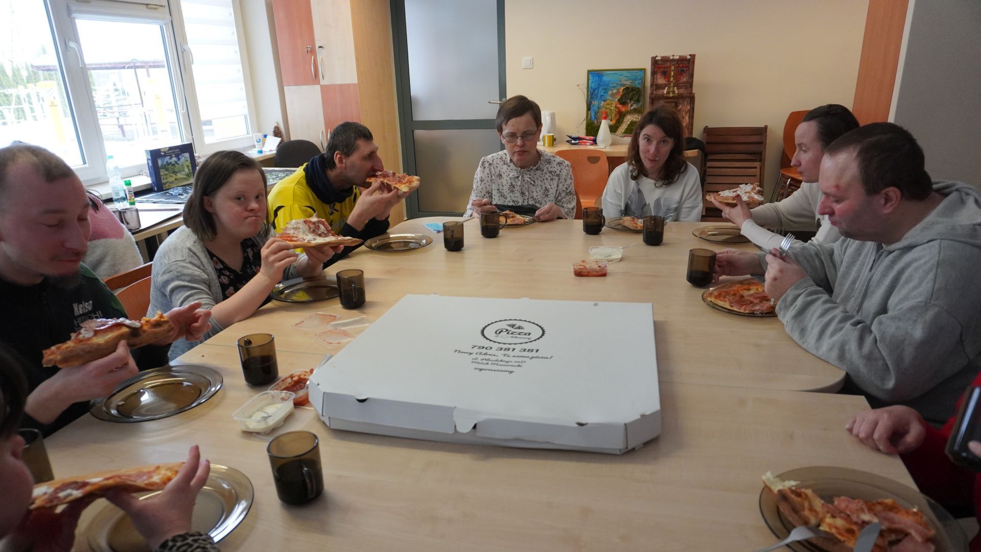 Uczestnicy przy wspólnym stole jedzą pizzę.