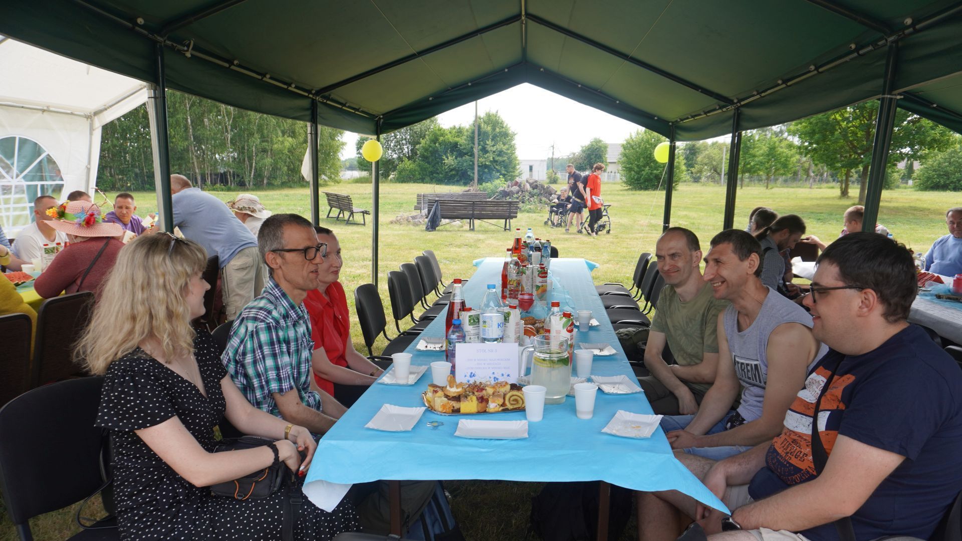 Uczestnicy wspólnie z terapeutą siedzą przy stole podczas Pikniku Rodzinnego ''Całkiem nowa bajka'' w DPS w Kątach.