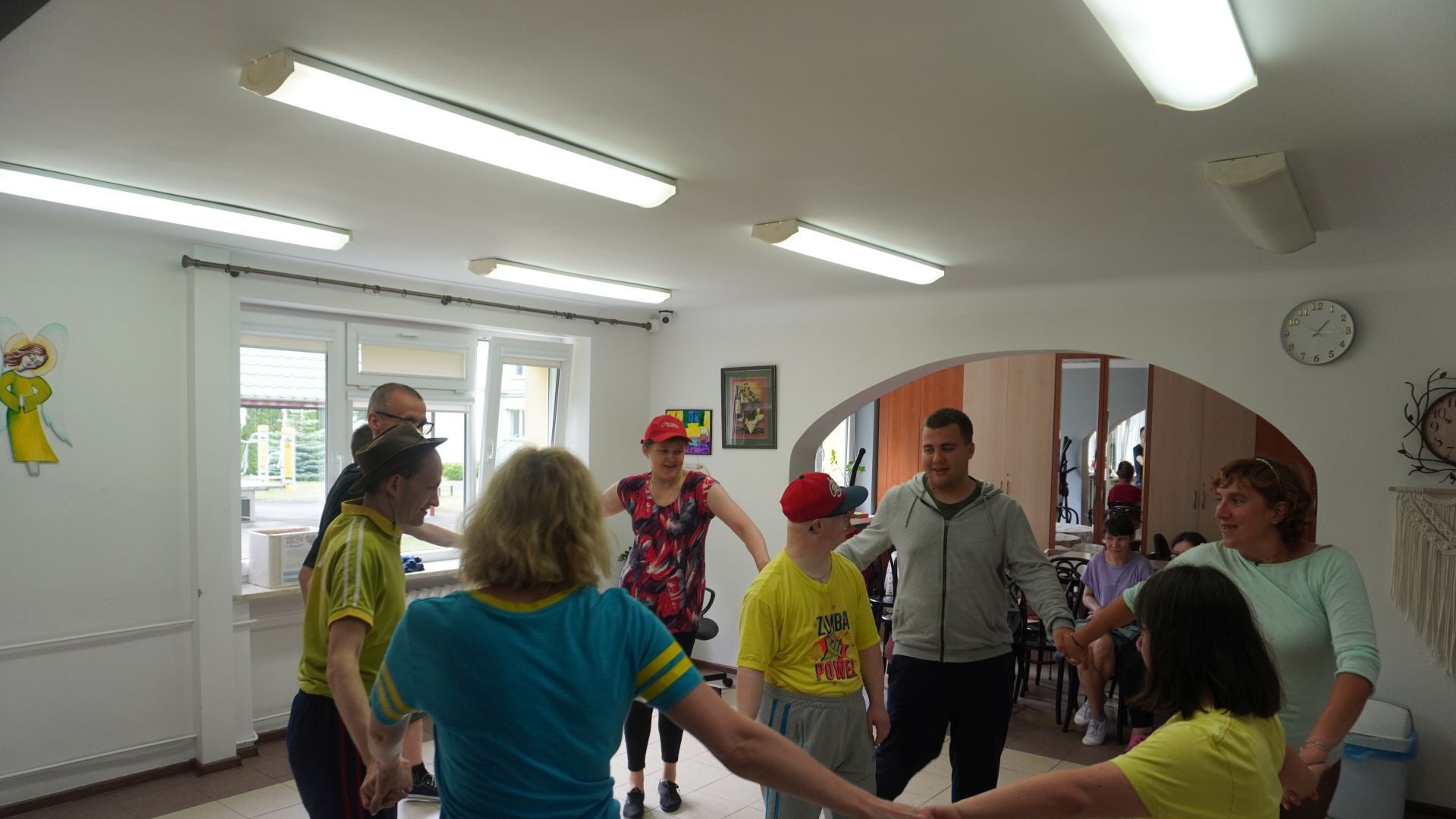 Uczestnicy wraz z terapeutą tańczą w kole podczas zajęć z elementami zumby, w środku stoi Uczestnik Mateusz. 