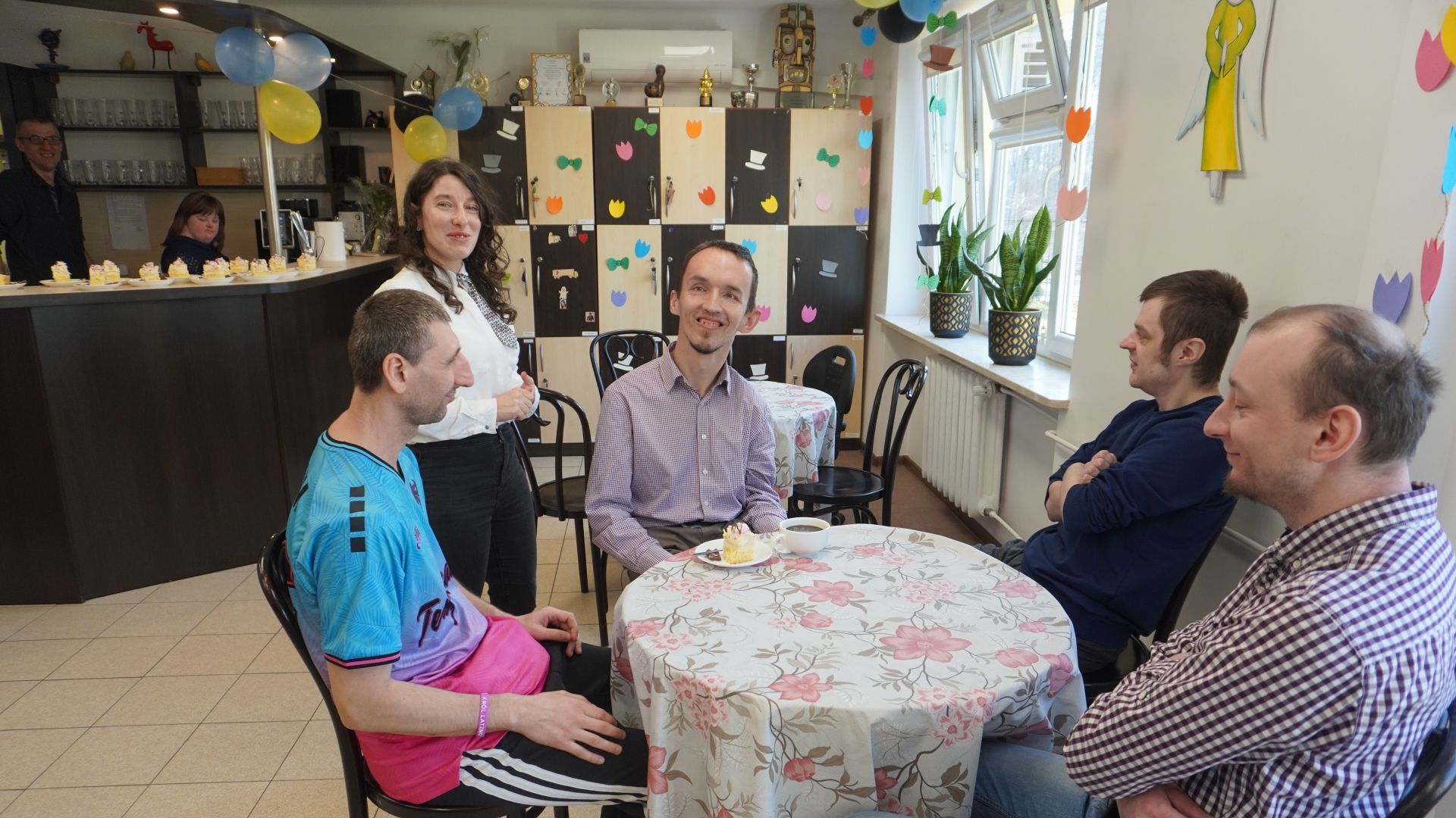 Justyna K. podaje kawę i ciasto do stolika, przy którym siedzą uczestnicy.