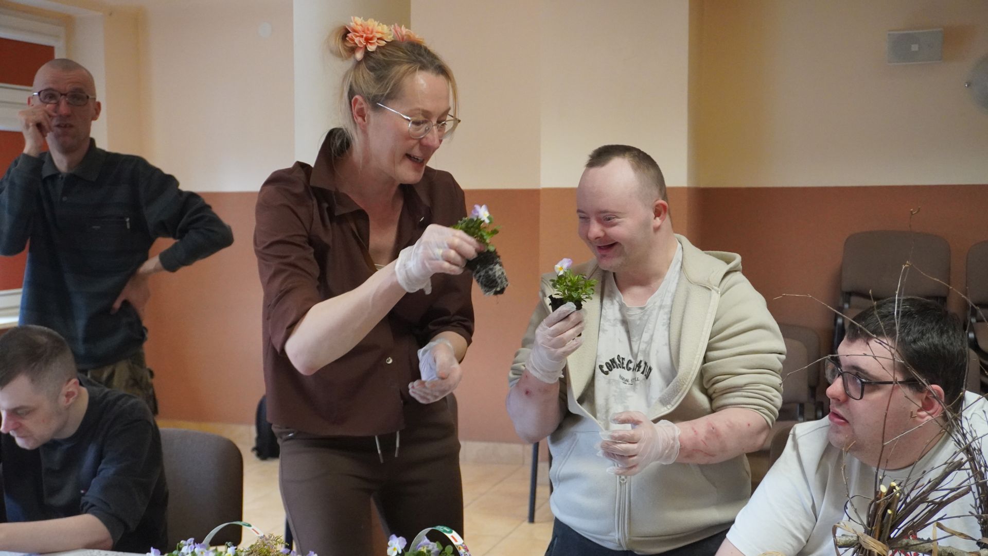 Uczestnik Darek podczas warsztatów wielkanocnych, z pomocą wolontariuszki, wkłada bratki do koszyków wykonanych z gałązek.