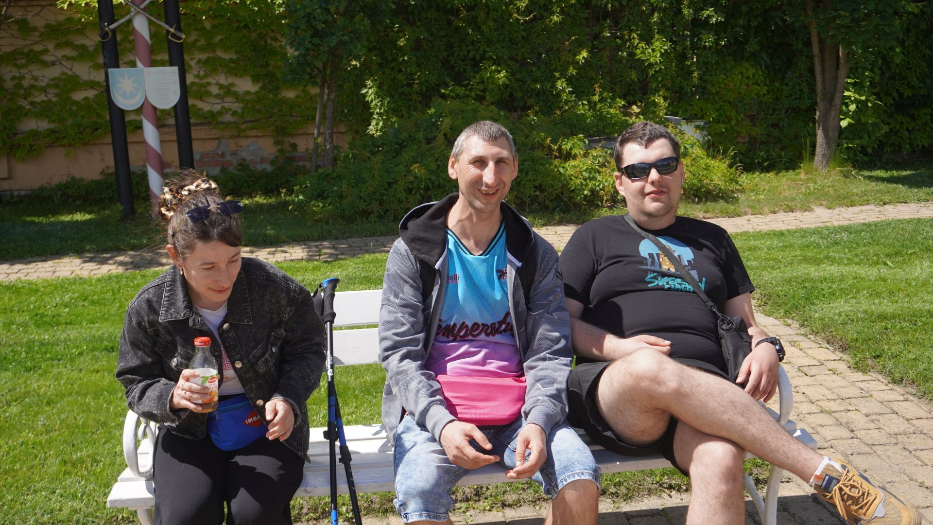Uczestnicy naszego Domu odpoczywają siedząc na ławce podczas spaceru z kijkami Nordic Walking.