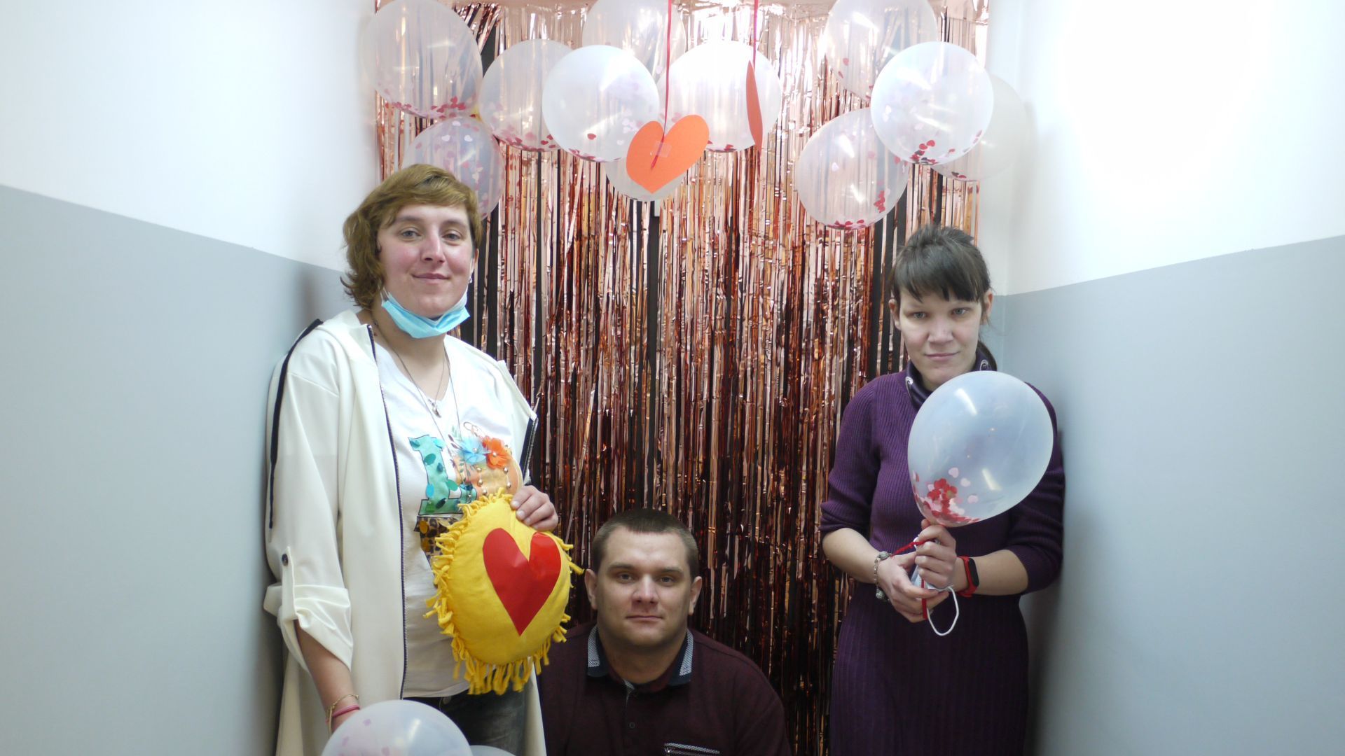 Uczestnicy z balonami i poduszką z serduszkiem na tle walentynkowej ścianki udekorowanej balonami i serpentynami. 