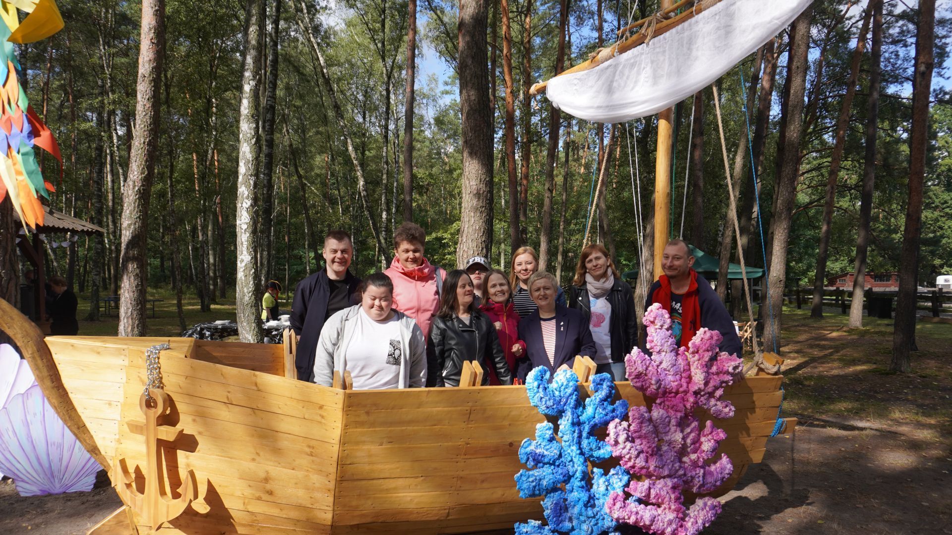 Uczestnicy wraz z panią dyrektor Justyną Juszczak i panią Kasią pozują do zdjęcia w drewnianej łodzi podczas imprezy plenerowej ''Morskie Opowieści'' w DPS ''Jedlina w Mieni.